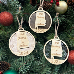 A festive ornament reading "First Christmas in Alabama" hangs on a holiday tree adorned with twinkling lights and evergreen branches. The ornament, shaped like the state of Alabama, features a warm, rustic design that captures the joy of the season.