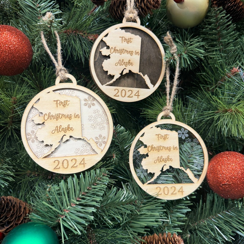 A festive ornament reading "First Christmas in Alaska" hangs on a holiday tree adorned with twinkling lights and evergreen branches. The ornament, shaped like the state of Alaska, features a warm, rustic design that captures the joy of the season.