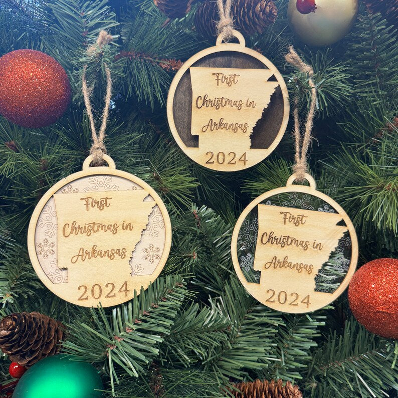 Rustic 'First Christmas in Arkansas' ornament shaped like the state of Arkansas, hanging on a glowing holiday tree with evergreen branches and twinkling lights. A charming keepsake to celebrate the warmth and traditions of the Natural State during the holiday season.