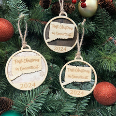 Connecticut-shaped 'First Christmas in Connecticut' ornament hanging on a beautifully decorated holiday tree with shimmering lights and lush branches. A timeless keepsake to celebrate a cozy and memorable first Christmas in the Constitution State.