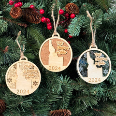 Idaho-shaped First Christmas in Idaho ornament hanging on a festive holiday tree with glowing lights and evergreen branches. A perfect keepsake to celebrate a cozy first Christmas in the Gem State, reflecting the beauty of its mountains and wilderness.