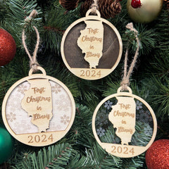 Illinois-shaped First Christmas in Illinois ornament hanging on a beautifully decorated holiday tree with twinkling lights and lush greenery. A wonderful keepsake to celebrate a special first Christmas in the Land of Lincoln, capturing the charm of its cities and countryside.