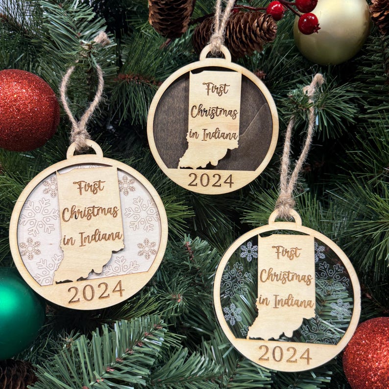 Indiana-shaped First Christmas in Indiana ornament hanging on a sparkling holiday tree with twinkling lights and evergreen branches. A perfect keepsake to celebrate a memorable first Christmas in the Hoosier State, filled with warmth and festive spirit.