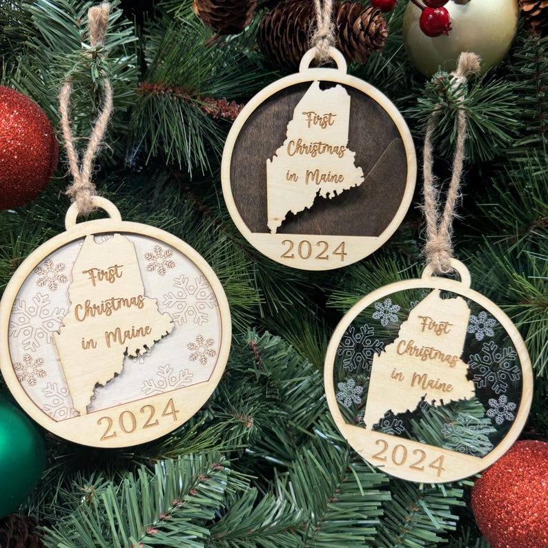 Maine-shaped First Christmas in Maine ornament hanging on a glowing holiday tree with twinkling lights and evergreen branches. A perfect keepsake to celebrate a cozy first Christmas in the Pine Tree State, reflecting its winter charm and serene landscapes.