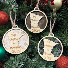 Minnesota-shaped First Christmas in Minnesota ornament hanging on a festive holiday tree with twinkling lights and evergreen branches. A charming keepsake to celebrate a cozy first Christmas in the Land of 10,000 Lakes, reflecting its winter wonderland and hearty holiday spirit.