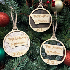 Montana-shaped First Christmas in Montana ornament hanging on a festive holiday tree with twinkling lights and evergreen branches. A perfect keepsake to celebrate a memorable first Christmas in the Treasure State, capturing its rugged beauty and winter charm.