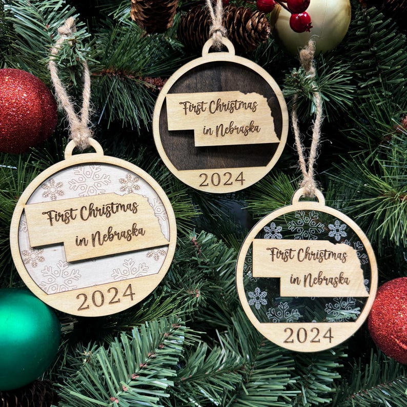 Nebraska-shaped First Christmas in Nebraska ornament hanging on a twinkling holiday tree with glowing lights and lush greenery. A perfect keepsake to celebrate a memorable first Christmas in the Cornhusker State, reflecting its wide-open skies and heartland spirit.