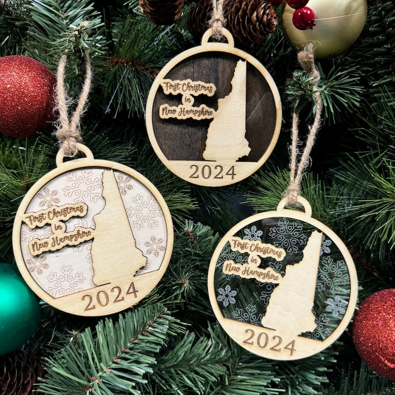 New Hampshire-shaped First Christmas in New Hampshire ornament hanging on a festive holiday tree with twinkling lights and evergreen branches. A charming keepsake to celebrate a memorable first Christmas in the Granite State, reflecting its mountainous landscapes and New England charm.