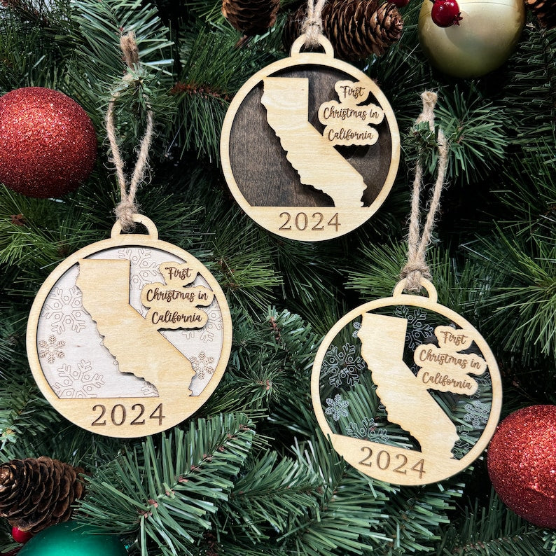 California-shaped 'First Christmas in California' ornament hanging on a radiant holiday tree with twinkling lights and lush greenery. A perfect way to celebrate a milestone holiday in the Golden State, capturing its iconic charm and festive spirit.