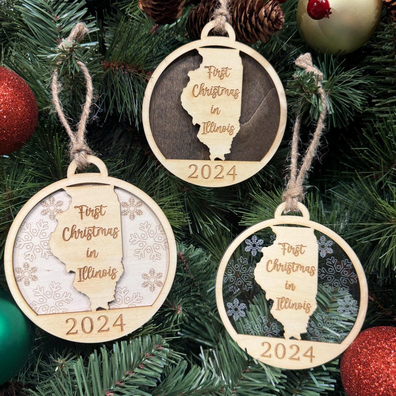 Illinois-shaped First Christmas in Illinois ornament hanging on a beautifully decorated holiday tree with twinkling lights and lush greenery. A wonderful keepsake to celebrate a special first Christmas in the Land of Lincoln, capturing the charm of its cities and countryside.