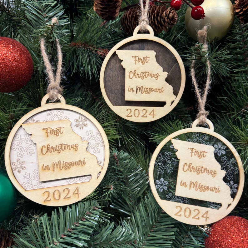 Missouri-shaped First Christmas in Missouri ornament hanging on a glowing holiday tree with twinkling lights and evergreen branches. A perfect keepsake to celebrate a special first Christmas in the Show-Me State, reflecting its diverse landscapes and holiday cheer.